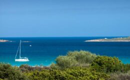 Sailboat anchored in calm turquoise waters with lush greenery in the foreground and distant islands under a clear blue sky.