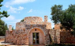 Stone structure with arched entrance surrounded by olive trees under a blue sky, showcasing traditional architecture.