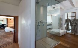 Modern bathroom design featuring a glass shower enclosure, wooden flooring, and a spacious layout. The image showcases a sleek, minimalist aesthetic with adjacent living areas and plush robes hanging nearby.