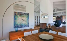 Modern dining area featuring a wooden table with white chairs, an arching floor lamp, and an abstract wall art piece. The space is illuminated with natural light and showcases a minimalist design aesthetic.