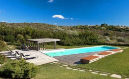 Modern outdoor swimming pool surrounded by lush greenery, featuring a wooden deck, lounge chairs, and a shaded pergola, ideal for relaxation and leisure activities.