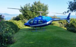 Blue helicopter parked on green lawn with trees and mountains in the background, showcasing a scenic outdoor setting.