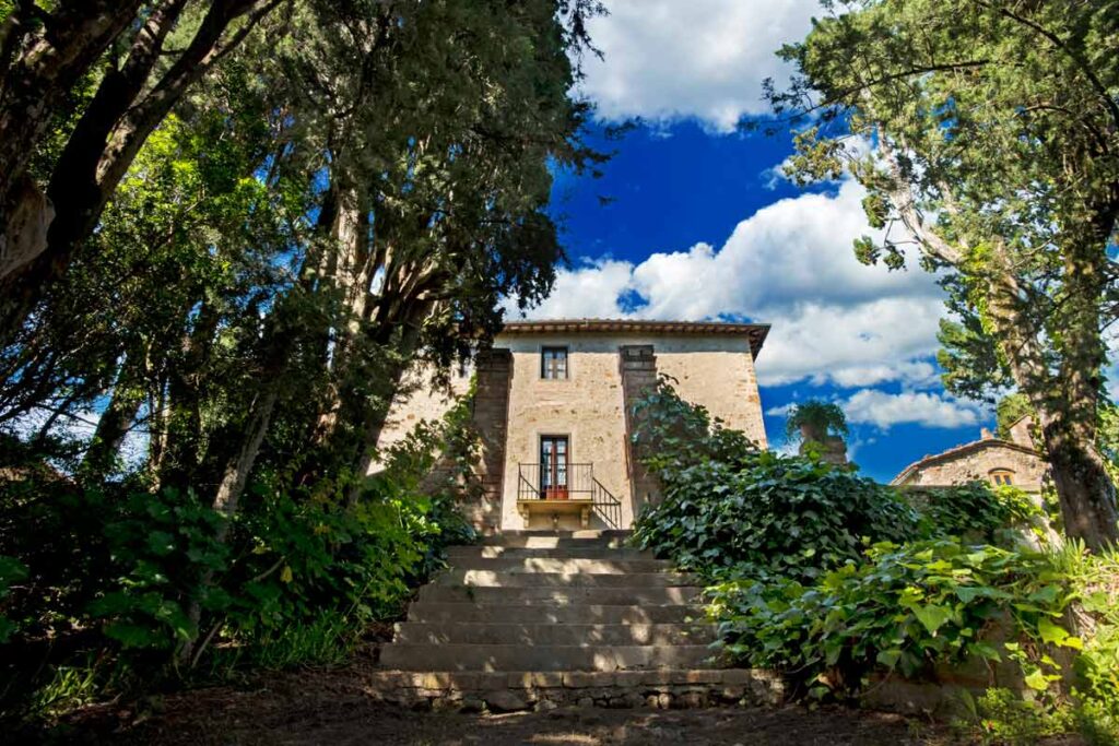 Stone house surrounded by lush greenery and trees, with a staircase leading up to the entrance under a bright blue sky with fluffy clouds. | Only on SopranoVillas