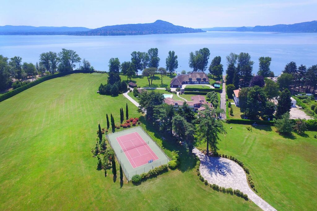 Aerial view of a luxurious lakeside property featuring a well-maintained lawn, a tennis court, and lush trees, with tranquil water and mountains in the background. Ideal for outdoor activities and relaxation. | Only on SopranoVillas