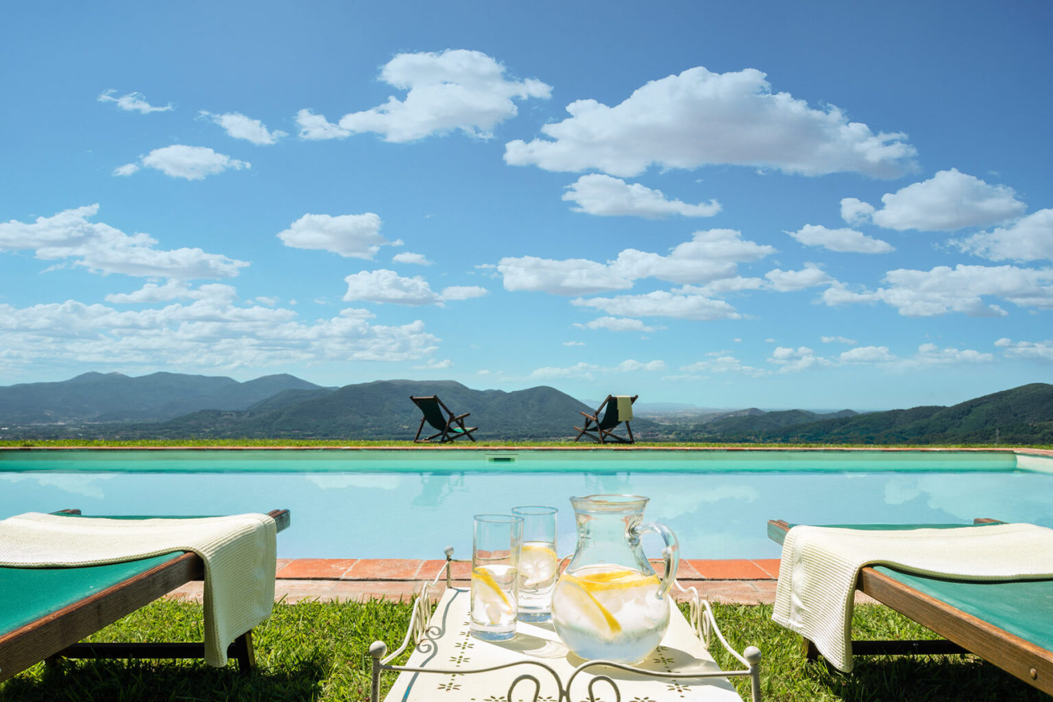 Serene outdoor poolside view with lounge chairs, a clear blue sky, and distant mountains, featuring a table with refreshing drinks and a pitcher, perfect for relaxation and leisure. | Only on SopranoVillas