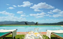 Serene outdoor poolside view with lounge chairs, a clear blue sky, and distant mountains, featuring a table with refreshing drinks and a pitcher, perfect for relaxation and leisure.