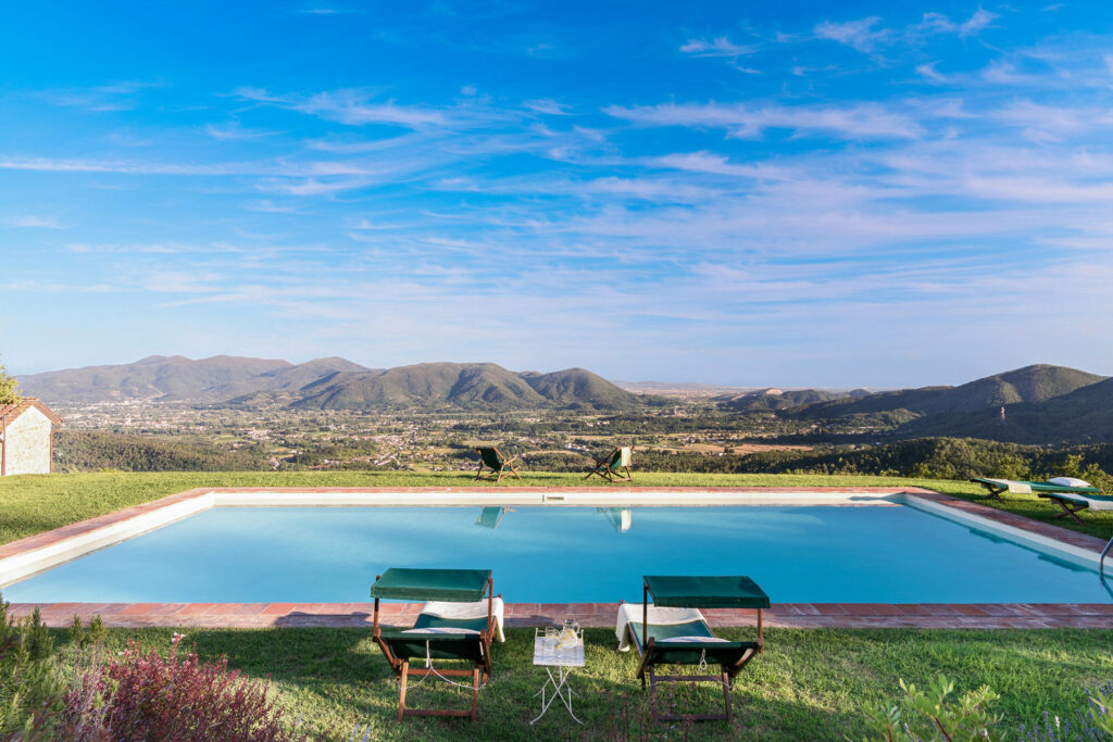 Scenic view of a tranquil swimming pool with two lounge chairs, surrounded by lush greenery and mountains under a clear blue sky. Perfect for relaxation and outdoor leisure. | Only on SopranoVillas