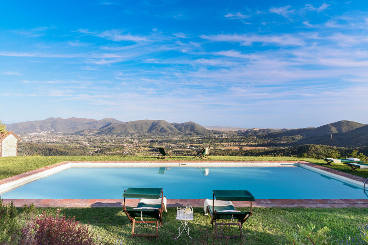 Scenic view of a tranquil swimming pool with two lounge chairs, surrounded by lush greenery and mountains under a clear blue sky. Perfect for relaxation and outdoor leisure. | Only on SopranoVillas