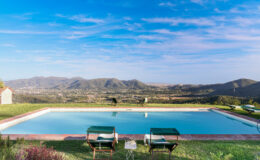 Scenic view of a tranquil swimming pool with two lounge chairs, surrounded by lush greenery and mountains under a clear blue sky. Perfect for relaxation and outdoor leisure.