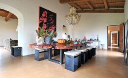 Spacious dining area featuring a large wooden table with decorative items, modern black stools, and a chandelier, complemented by a vibrant artwork on the wall and natural light from arched doorways.