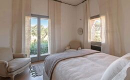 Cozy bedroom featuring a queen-sized bed with a light beige blanket, soft armchair, large windows with sheer curtains, and a view of greenery outside, creating a serene and inviting atmosphere.