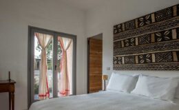 Cozy bedroom featuring a comfortable bed with white linens, a decorative patterned wall hanging, and large windows with flowing curtains that brighten the space.