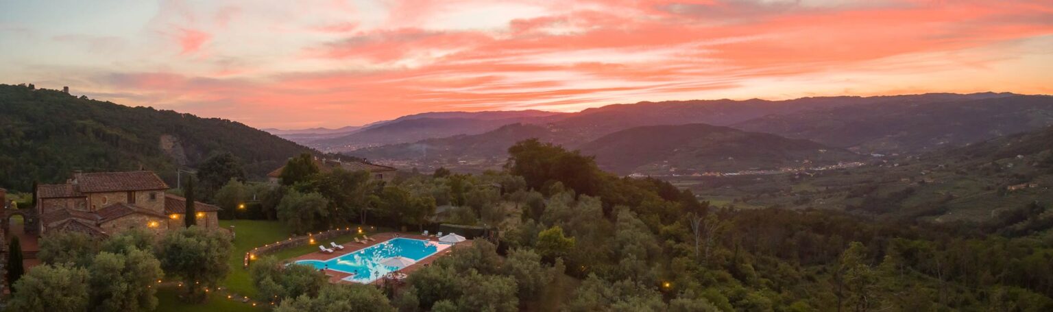 Aerial view of a serene swimming pool surrounded by lush greenery at sunset, with vibrant pink and orange skies and rolling hills in the background. | Only on SopranoVillas