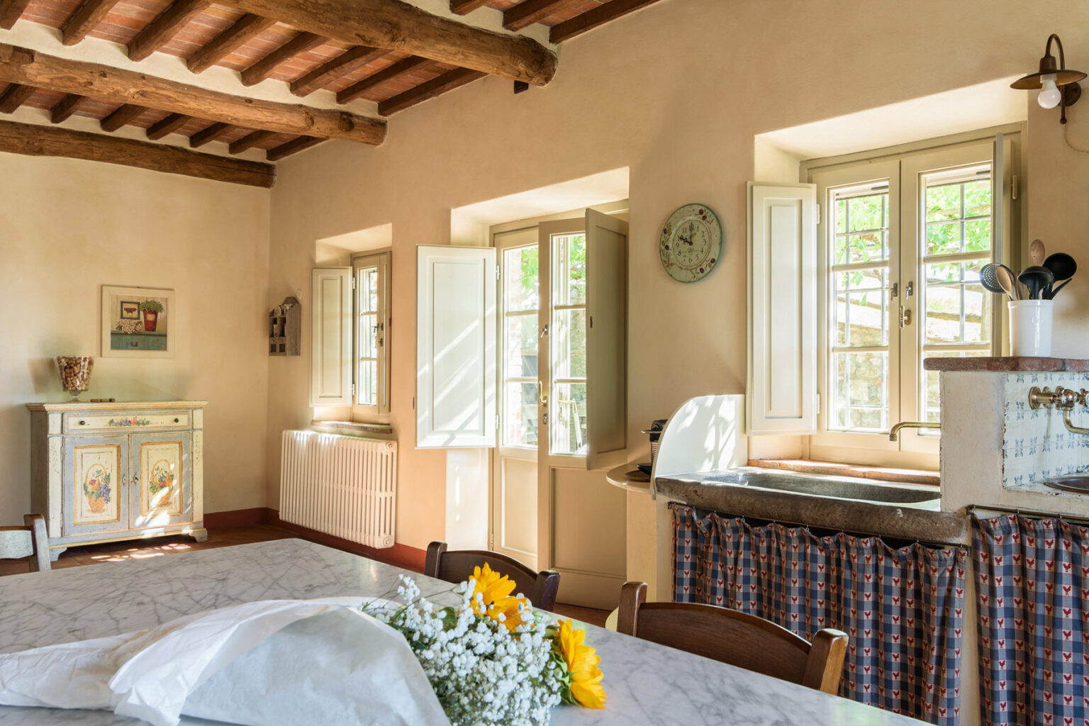 Bright and airy kitchen interior featuring wooden beams, large open windows, and a rustic dining table adorned with sunflowers. The space includes a vintage sideboard and a practical sink area, creating a warm and inviting atmosphere. | Only on SopranoVillas