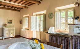 Bright and airy kitchen interior featuring wooden beams, large open windows, and a rustic dining table adorned with sunflowers. The space includes a vintage sideboard and a practical sink area, creating a warm and inviting atmosphere.