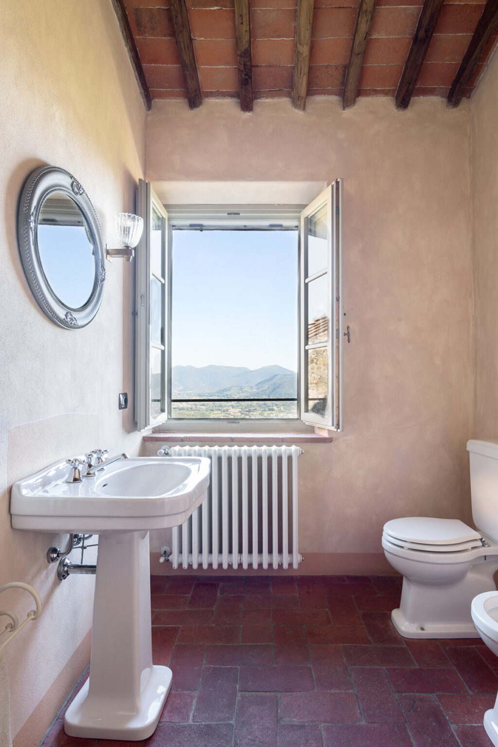 Modern bathroom with a scenic view, featuring a sink, mirror, and window overlooking mountains. Natural light enhances the warm, earthy tones of the walls. | Only on SopranoVillas