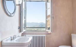Modern bathroom with a scenic view, featuring a sink, mirror, and window overlooking mountains. Natural light enhances the warm, earthy tones of the walls.