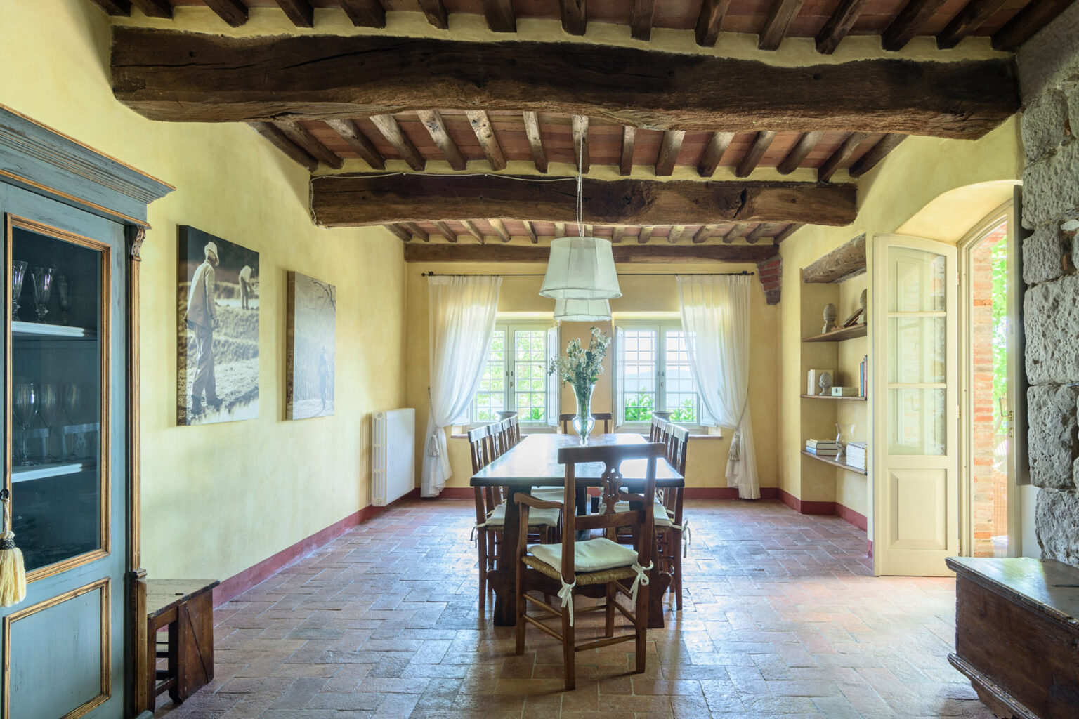 Spacious dining room with rustic wooden beams, a large wooden table surrounded by chairs, and natural light streaming through large windows, featuring a cozy and inviting atmosphere. | Only on SopranoVillas