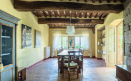 Spacious dining room with rustic wooden beams, a large wooden table surrounded by chairs, and natural light streaming through large windows, featuring a cozy and inviting atmosphere.