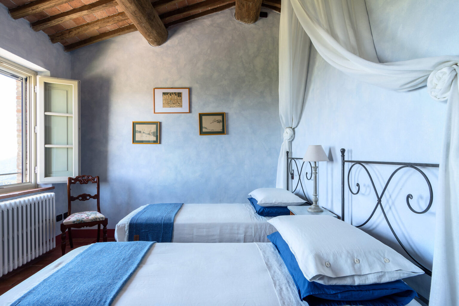Bright and airy bedroom featuring two twin beds with blue accents, a wooden chair, and framed artwork on the walls. The room has a rustic charm with exposed wooden beams and a large window allowing natural light to fill the space. Ideal for a cozy getaway or guest accommodation. | Only on SopranoVillas