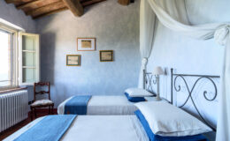 Bright and airy bedroom featuring two twin beds with blue accents, a wooden chair, and framed artwork on the walls. The room has a rustic charm with exposed wooden beams and a large window allowing natural light to fill the space. Ideal for a cozy getaway or guest accommodation.