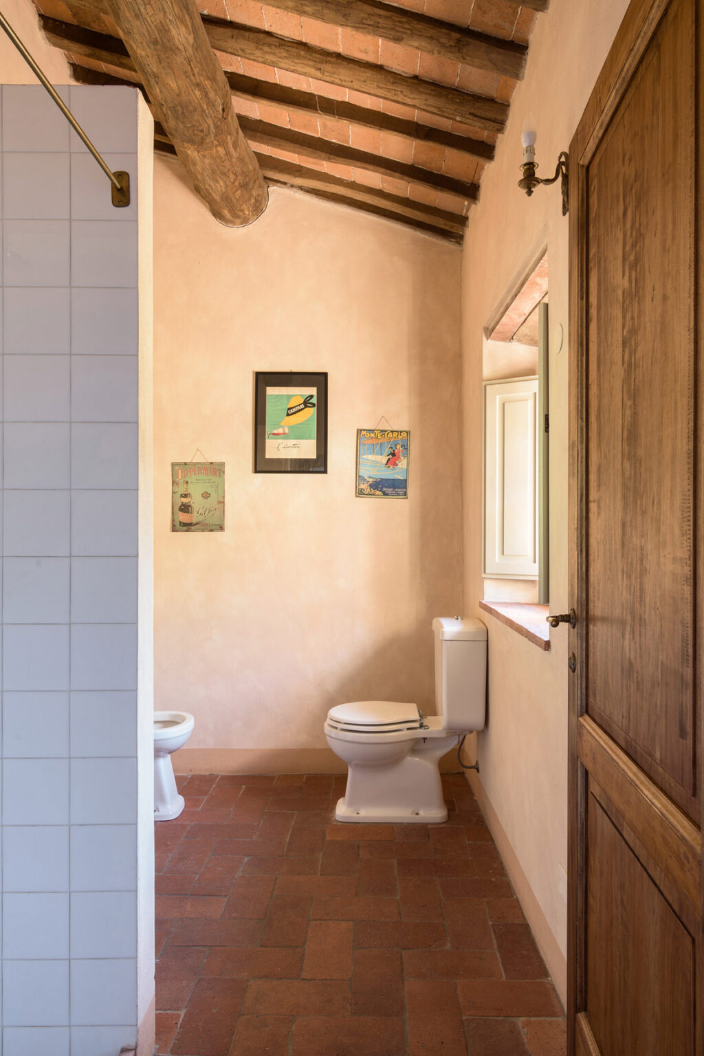Cozy bathroom interior featuring a white toilet, rustic wooden door, and framed art on a textured peach wall, with a window allowing natural light. | Only on SopranoVillas