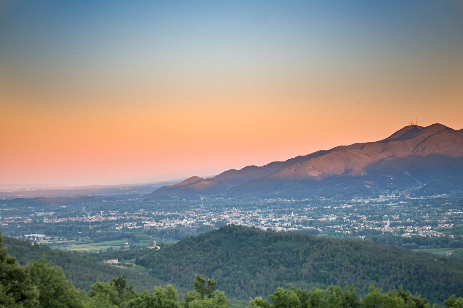 Scenic view of mountains at sunset, showcasing a vibrant sky with hues of orange and blue, overlooking a valley and a town below. | Only on SopranoVillas