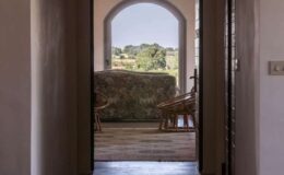 Interior view of a doorway leading to a scenic outdoor landscape, featuring a cozy seating area with floral-patterned upholstery and wooden chairs, framed by an arched entrance.