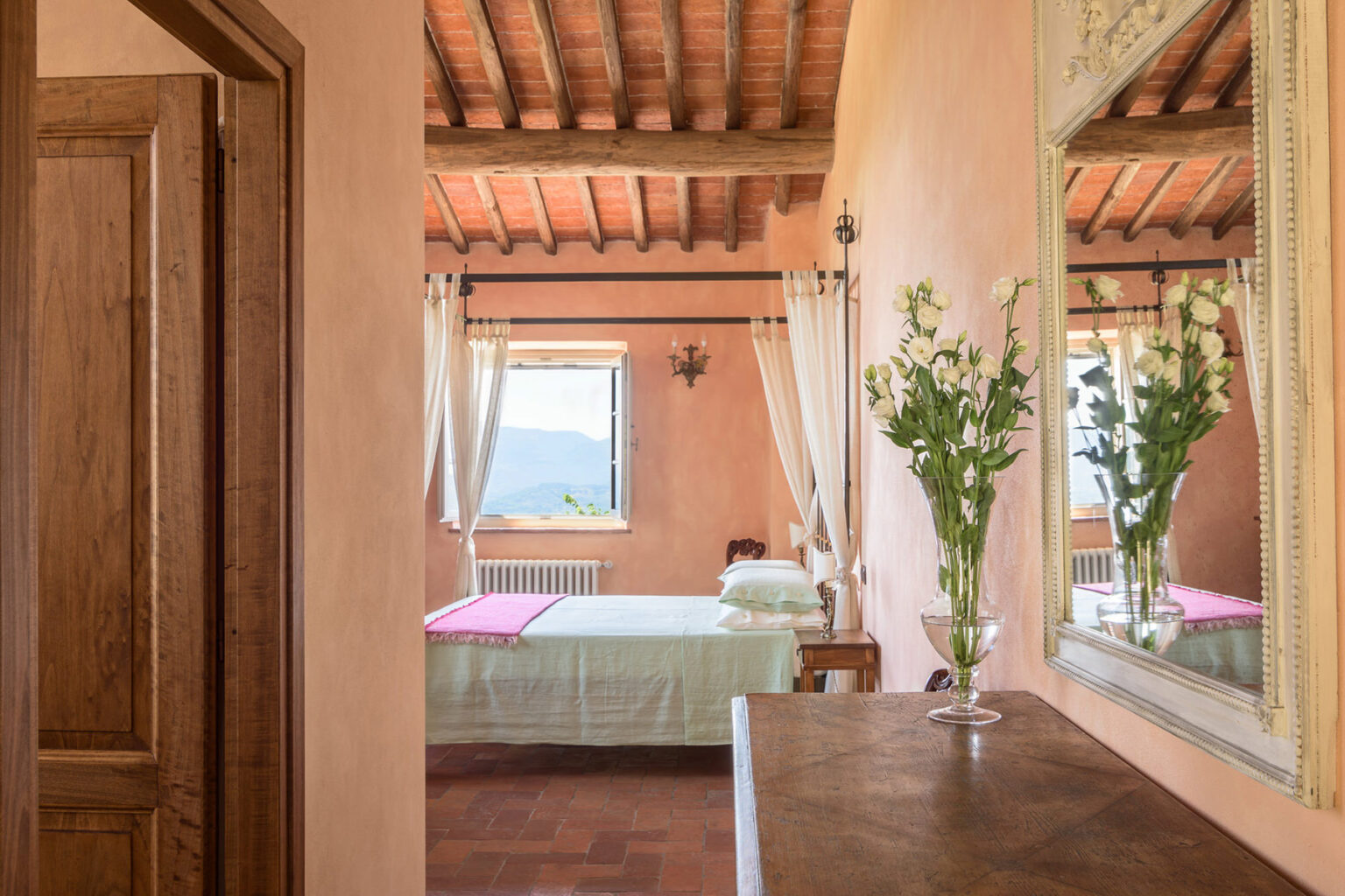 Cozy bedroom interior featuring rustic wooden beams, warm peach-colored walls, a large mirror, and a vase of fresh flowers on a dresser, with natural light streaming through a window. | Only on SopranoVillas