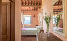 Cozy bedroom interior featuring rustic wooden beams, warm peach-colored walls, a large mirror, and a vase of fresh flowers on a dresser, with natural light streaming through a window.
