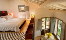 Cozy bedroom interior featuring a queen-sized bed, wooden furniture, and large windows with natural light. The room has a stylish checkered floor and a warm ambiance, perfect for relaxation.