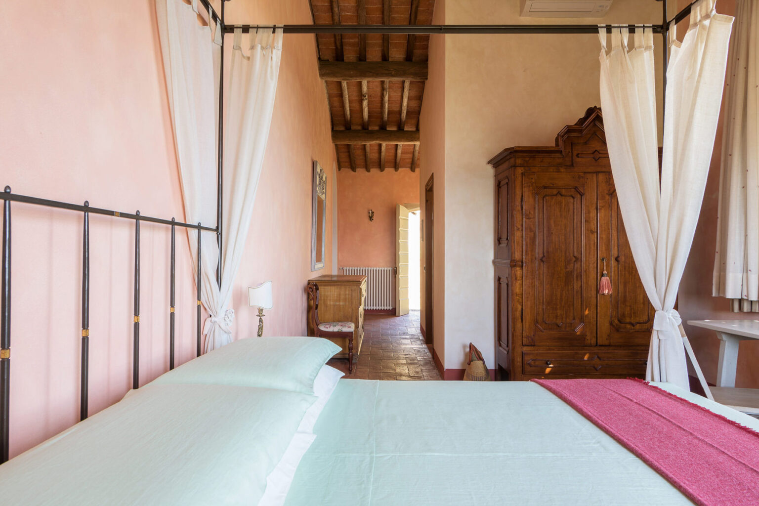 Cozy bedroom featuring a wrought iron bed with light green linens, rustic wooden furniture, and soft natural light illuminating the warm peach walls. The room includes a vintage wardrobe and sheer curtains, creating a tranquil atmosphere. | Only on SopranoVillas