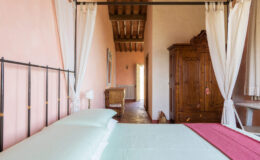 Cozy bedroom featuring a wrought iron bed with light green linens, rustic wooden furniture, and soft natural light illuminating the warm peach walls. The room includes a vintage wardrobe and sheer curtains, creating a tranquil atmosphere.