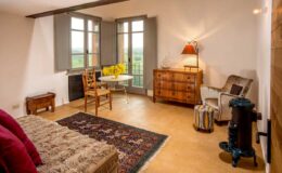 Cozy bedroom interior featuring a plush bed with decorative pillows, a wooden dresser, a comfortable armchair, and a small dining table by large windows with scenic views. The room is illuminated by a stylish lamp and has rustic decor elements, creating a warm and inviting atmosphere.