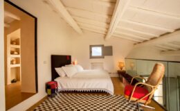 Modern loft bedroom featuring a cozy double bed, stylish red armchair, and large windows that allow natural light, complemented by a checkered rug and minimalist decor.