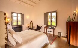 Cozy bedroom featuring a queen-sized bed with white linens, large windows with green shutters, a vintage mirror, a stylish chair, and a wooden dresser, creating a warm and inviting atmosphere.