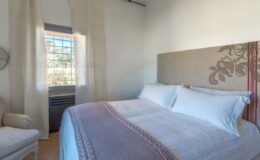 Cozy bedroom featuring a comfortable double bed with white linens, decorative throw, and natural light streaming through a window, creating a serene atmosphere.