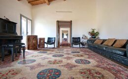 Spacious living room featuring a vintage piano, black leather seating, and a decorative area rug with intricate patterns, complemented by natural light from large windows.