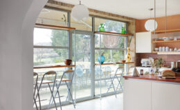 Modern kitchen interior featuring large glass doors, wooden shelves with decorative items, and a minimalist design. The space includes a sleek countertop and folding chairs, creating a bright and inviting atmosphere.