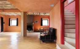 Spacious living area with orange walls, wooden ceiling beams, and a staircase leading to the upper level, featuring a cozy seating arrangement and a fireplace.