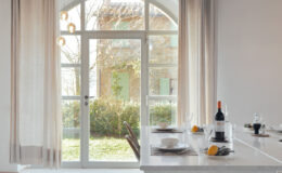 Modern dining area with a view of a garden through large glass doors, featuring a white marble table set with tableware, a bottle of wine, and fresh fruits. Natural light illuminates the space, enhancing its elegant ambiance.