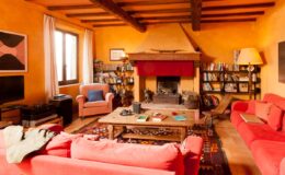 Cozy living room featuring orange walls, wooden beams, red sofas, a central coffee table, and a fireplace, with shelves filled with books and decorative items. Natural light streams in through large windows, creating a warm and inviting atmosphere.