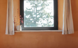 Cozy interior view featuring a large window with black frame, white curtains, and a small potted plant on a stone windowsill, set against warm orange walls and a backdrop of trees outside.