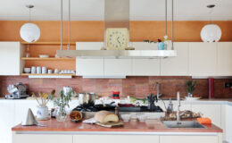 Modern kitchen interior with white cabinets, a central island, and a mixture of cooking utensils and fresh ingredients. Features a stylish clock, hanging light fixtures, and a warm orange accent wall, creating a cozy and inviting cooking space.