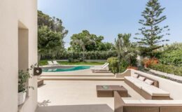 Modern outdoor patio with a comfortable seating area overlooking a swimming pool surrounded by lush greenery and trees, perfect for relaxation and entertaining.
