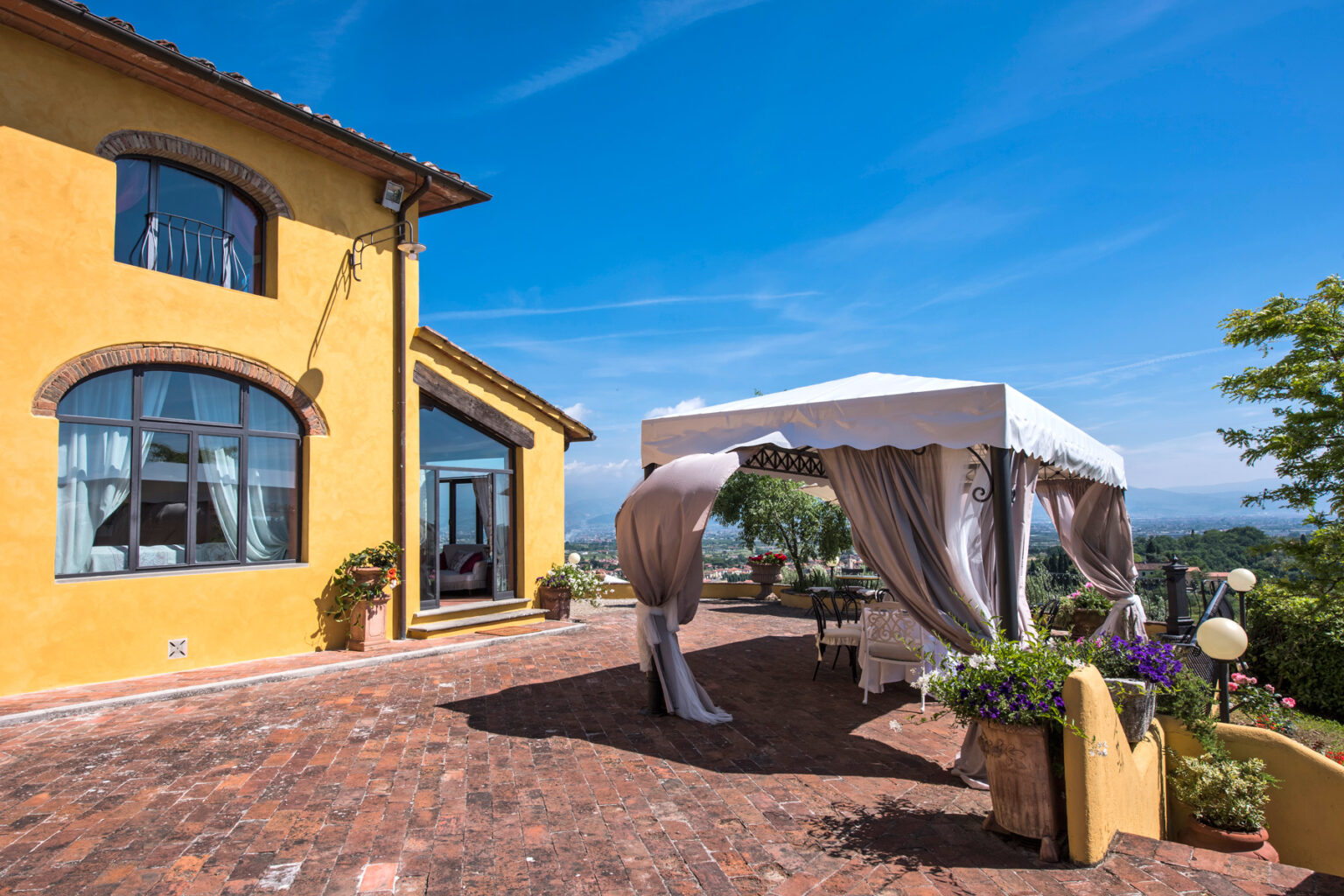 Charming yellow villa with a spacious terrace featuring a white canopy, surrounded by lush greenery and scenic views under a clear blue sky. Ideal for outdoor events and gatherings. | Only on SopranoVillas