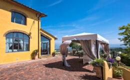 Charming yellow villa with a spacious terrace featuring a white canopy, surrounded by lush greenery and scenic views under a clear blue sky. Ideal for outdoor events and gatherings.