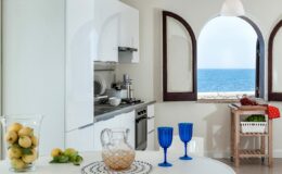 Bright coastal kitchen with sea view, featuring white cabinetry, a round dining table with blue glassware, and a bowl of lemons, creating a fresh and inviting atmosphere.