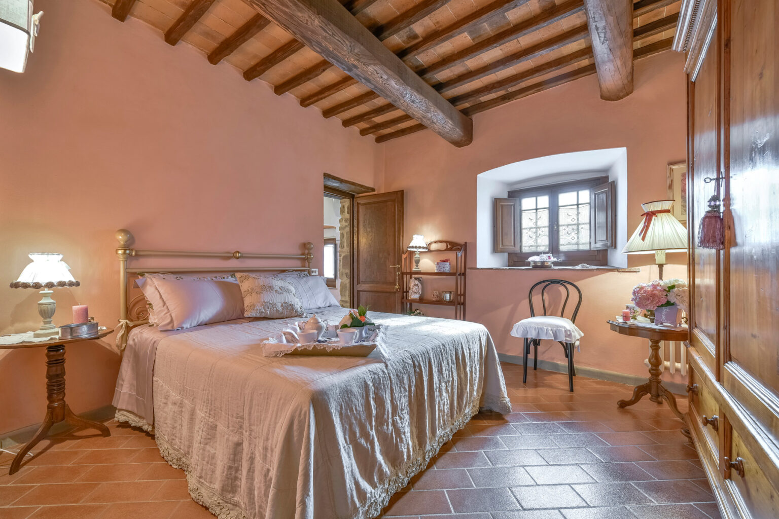 Cozy bedroom featuring a wooden beam ceiling, warm terracotta walls, and a comfortable double bed with soft linens. A bedside table with a lamp and a decorative chair enhance the inviting atmosphere. Natural light filters through the window, creating a serene ambiance. | Only on SopranoVillas