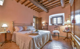 Cozy bedroom featuring a wooden beam ceiling, warm terracotta walls, and a comfortable double bed with soft linens. A bedside table with a lamp and a decorative chair enhance the inviting atmosphere. Natural light filters through the window, creating a serene ambiance.